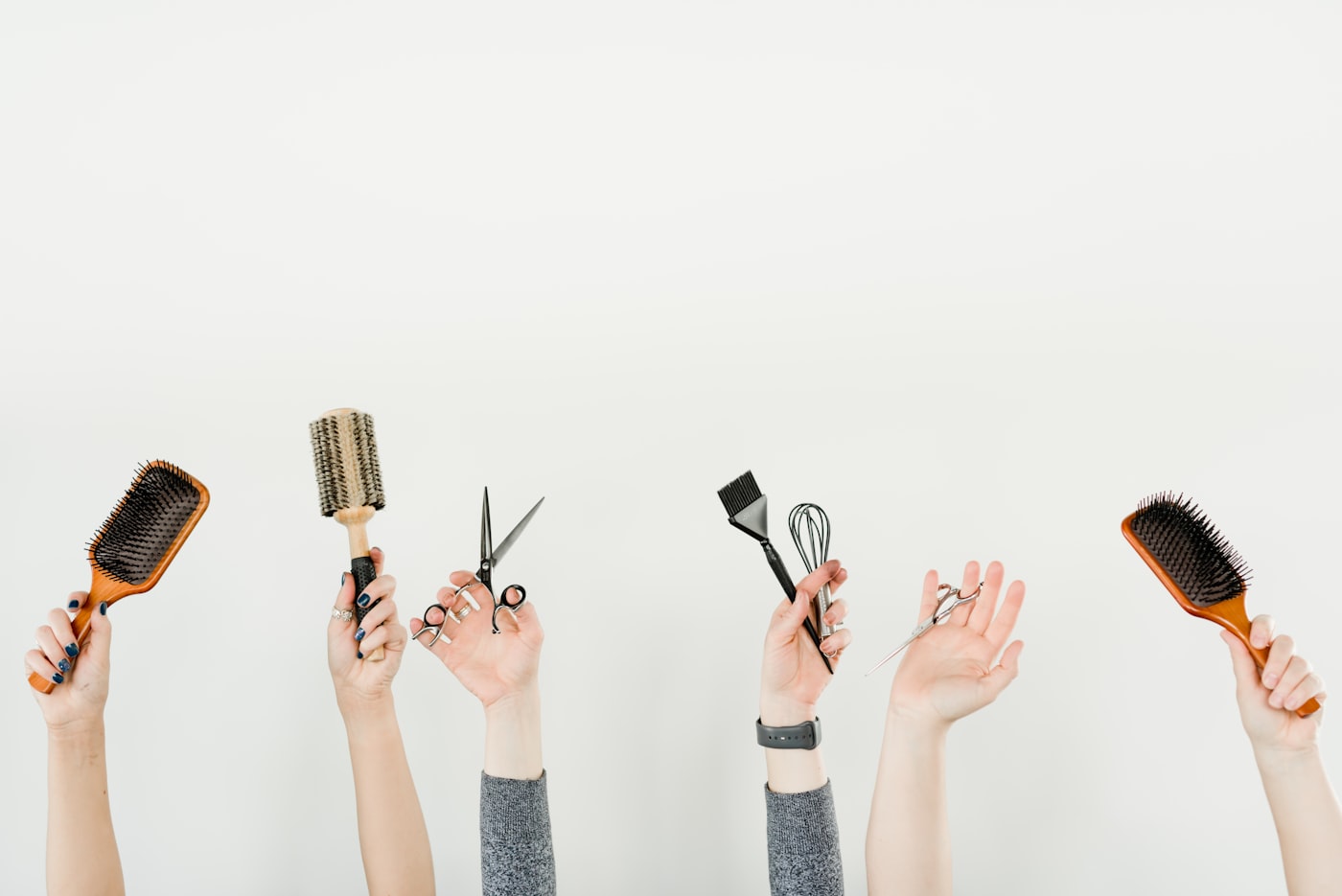 Four hands holding brushes, shears and a comb arranged in a row against a plain wall